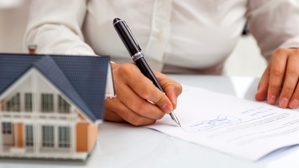 Persona firmando documentos relacionados con una propiedad junto a una maqueta de vivienda 


