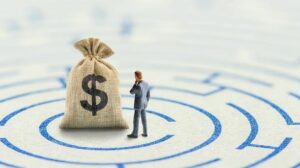 Empresario frente a un saco de dinero sobre una ruta circular, que representa la búsqueda de financiamiento para empresas para obtener capital, mejorar la liquidez y avanzar hacia nuevos objetivos.