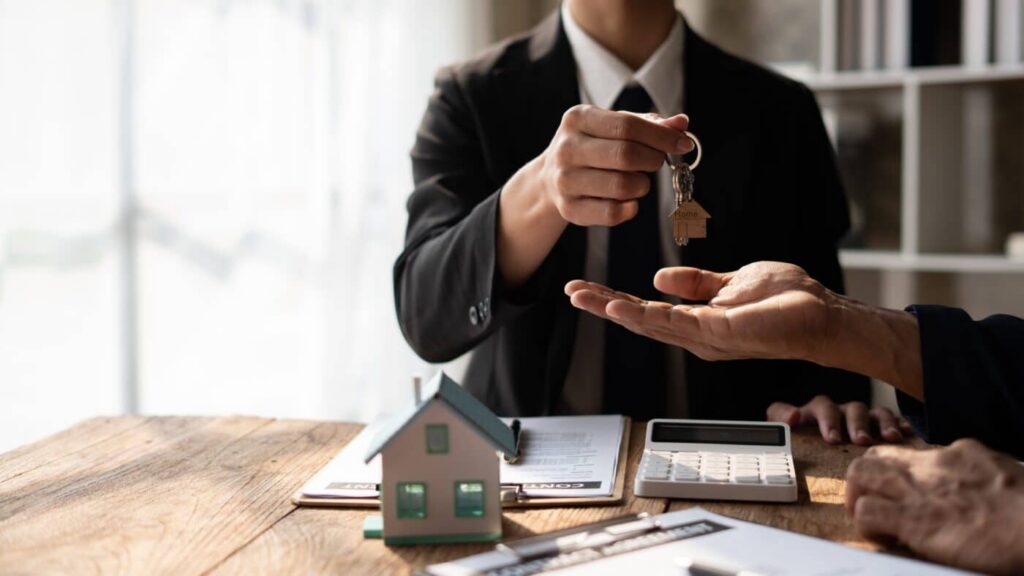 Persona entregando las llaves de una vivienda durante una operación de arrendamiento, con documentos y una calculadora que representan las condiciones financieras del contrato. 